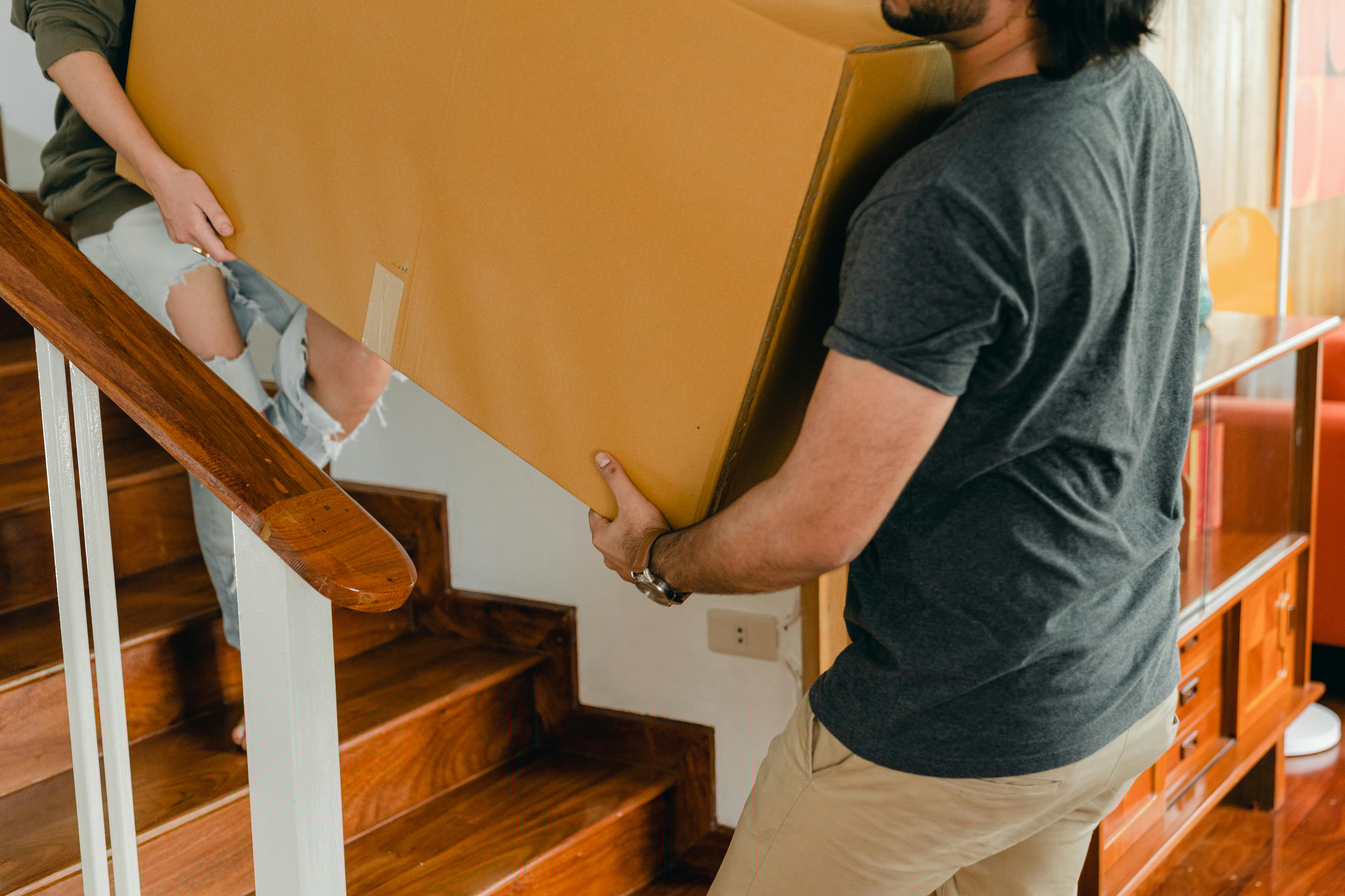 Two individuals carrying a large cardboard box up a wooden staircase inside a house.