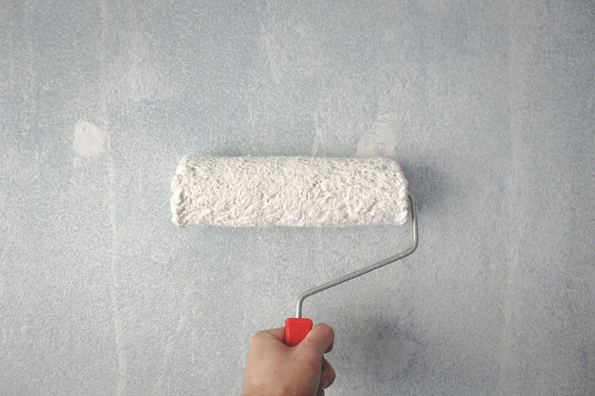 Hand holding a paint roller with white paint, painting a light gray wall.